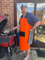 Man wearing an orange apron standing next to a barbecue grill outdoors.