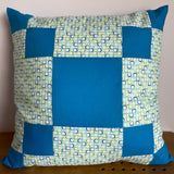 A square patchwork cushion, perfect for those taking a patchwork & quilting for beginners course, features solid blue squares and patterned fabric with blue and green circles on a white background, placed against a light wall.